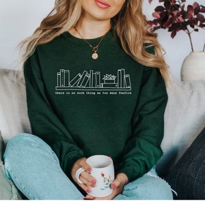 May include: A dark green sweatshirt with a white graphic of a bookshelf and the text "there is no such thing as too many fanfics." The person is holding a white mug with floral designs.