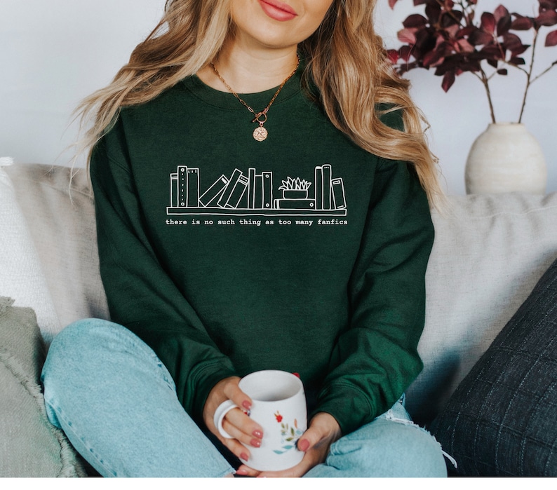 May include: A dark green sweatshirt with a white graphic of a bookshelf and the text "there is no such thing as too many fanfics." The person is holding a white mug with floral designs.