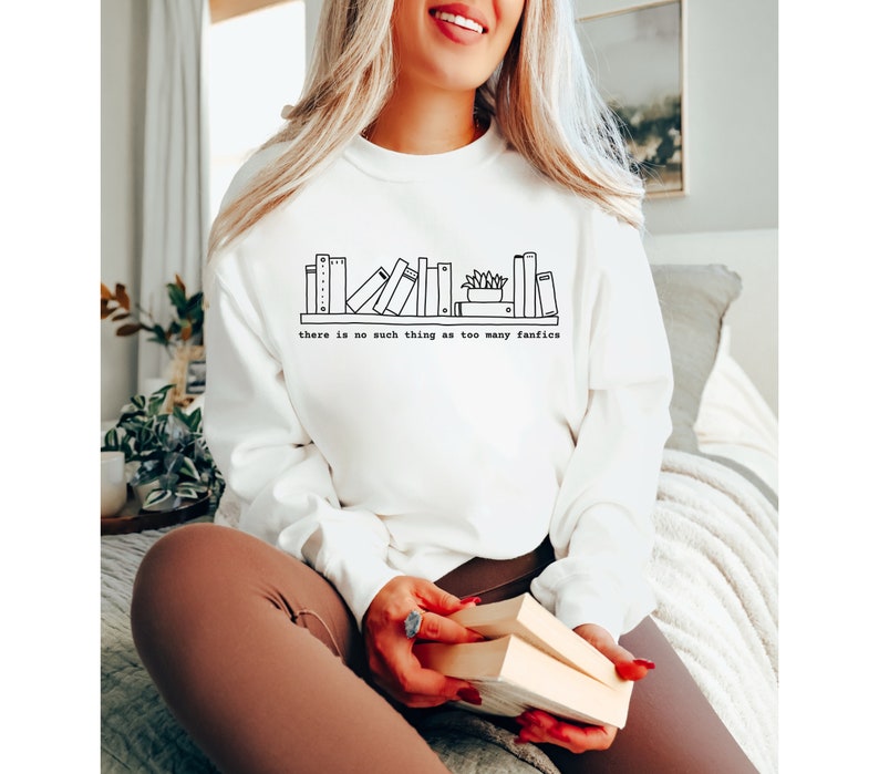 May include: White long-sleeve sweatshirt with a black line drawing of a bookshelf. The text "there is no such thing as too many fanfics" is printed below the bookshelf graphic. The sweatshirt is a casual style.