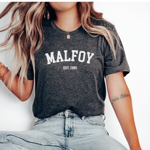 May include: A dark grey t-shirt with white text that reads "MALFOY EST. 1980".
