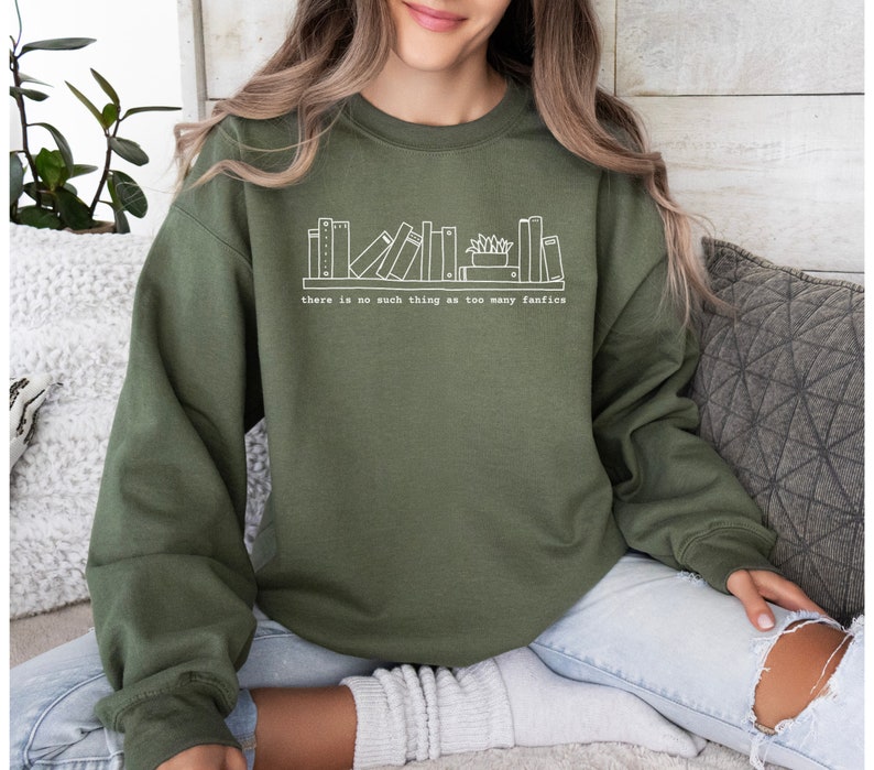 May include: An olive green sweatshirt with a white graphic of a bookshelf and the text "there is no such thing as too many fanfics". The sweatshirt is a long-sleeved, crew neck style.