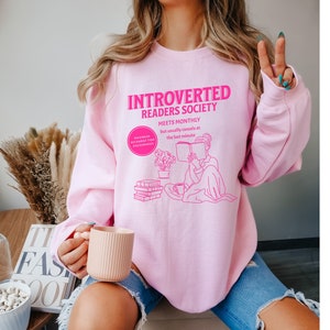 May include: Pink sweatshirt with the text "Introverted Readers Society Meets Monthly but usually cancels at the last minute" in pink. The graphic shows a person reading a book with a cup of tea and a stack of books.