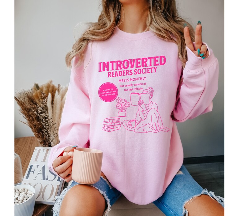May include: Pink sweatshirt with the text "Introverted Readers Society Meets Monthly but usually cancels at the last minute" in pink. The graphic shows a person reading a book with a cup of tea and a stack of books.