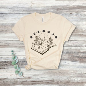 May include: A cream-colored t-shirt with a black and white graphic of an open book with mushrooms, ferns, and a crescent moon. The book is surrounded by a circle of moons in different phases.