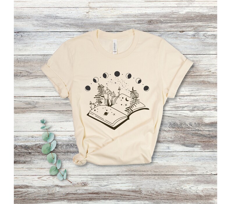 May include: A cream-colored t-shirt with a black and white graphic of an open book with mushrooms, ferns, and a crescent moon. The book is surrounded by a circle of moons in different phases.