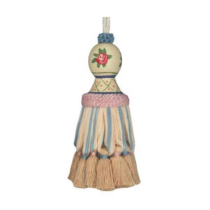 Puede incluir: Una borla decorativa con una cuenta de madera redonda pintada con una rosa roja y hojas verdes, rematada con un nudo azul. La borla incluye una banda texturizada rosa sobre una falda de hilos de tela alternos en color crema y azul con flecos.