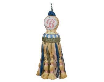 May include: A yellow and blue tassel with a painted round top featuring the letter "B" in pink. The top is decorated with small white flowers and blue stripes.