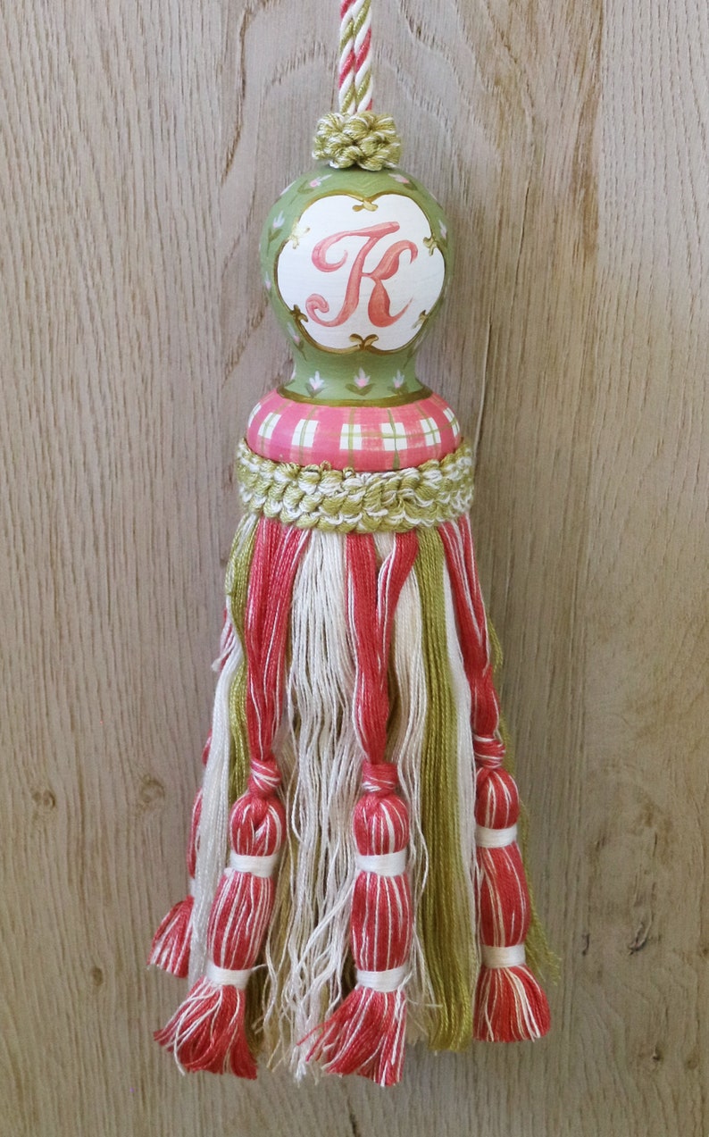 Puede incluir: Un borl&oacute;n verde y rosa con una parte superior de madera que presenta la letra "K" en rosa. El borl&oacute;n est&aacute; hecho de hilo verde, blanco y rosa.