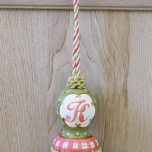 Puede incluir: Un borl&oacute;n verde y rosa con un c&iacute;rculo blanco que presenta la letra "K" en rosa. El borl&oacute;n tiene un patr&oacute;n de cuadros blancos y rosas y un cord&oacute;n a rayas verde y blanco.