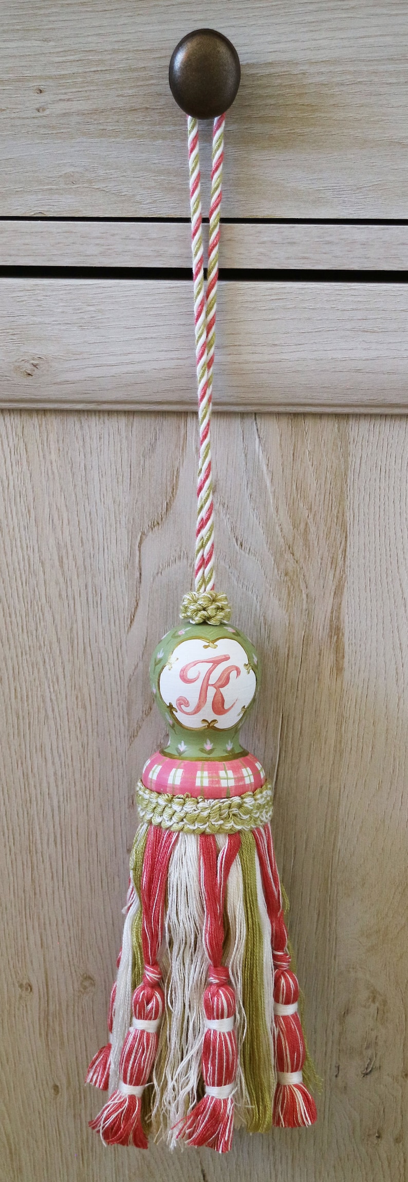 Puede incluir: Un borl&oacute;n verde y rosa con un c&iacute;rculo blanco que presenta la letra "K" en rosa. El borl&oacute;n tiene un patr&oacute;n de cuadros blancos y rosas y un cord&oacute;n a rayas verde y blanco.