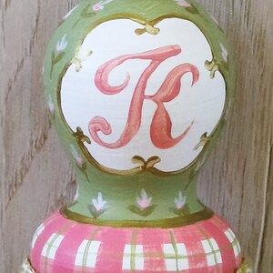 Puede incluir: Un adorno pintado a mano de color verde y rosa con fondo blanco y la letra "K" en rosa. El adorno tiene un patr&oacute;n de cuadros en la parte inferior y una borla verde y blanca en la parte superior.