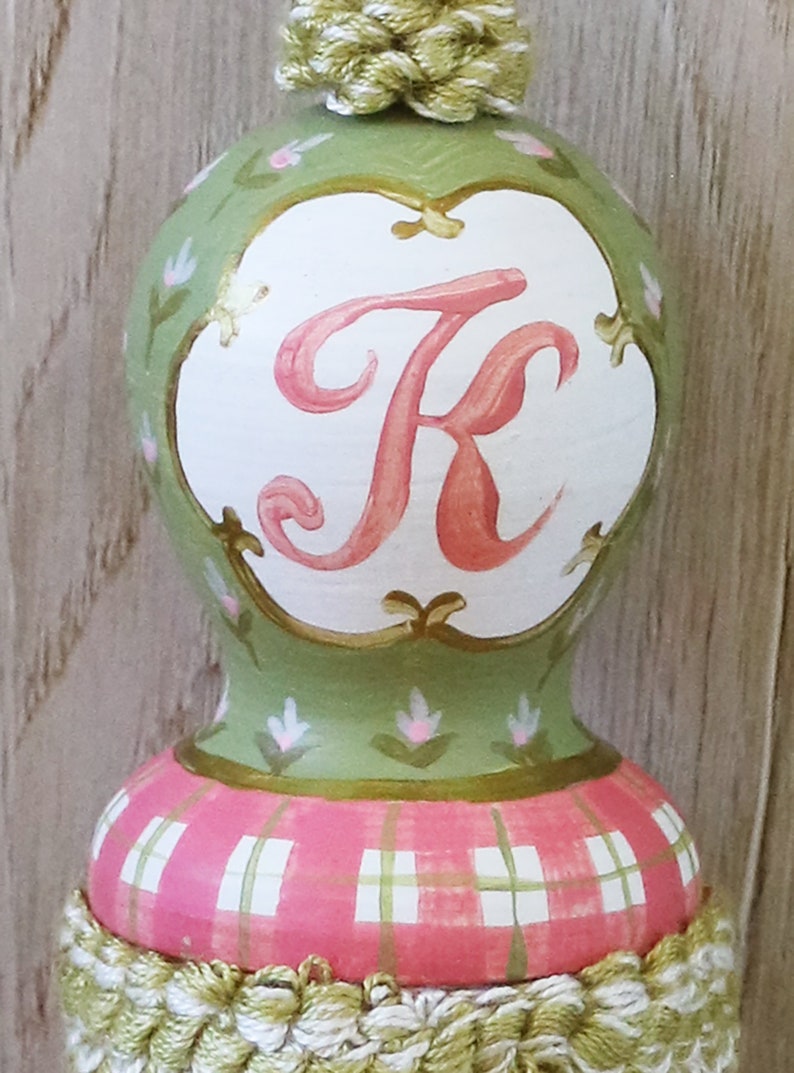 Puede incluir: Un adorno pintado a mano de color verde y rosa con fondo blanco y la letra "K" en rosa. El adorno tiene un patr&oacute;n de cuadros en la parte inferior y una borla verde y blanca en la parte superior.