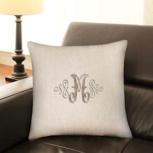 May include: A square, off-white decorative pillow with a gray embroidered monogram letter "M" in a decorative script. The pillow is resting on a dark brown leather couch. A lamp with a beige shade is in the background.