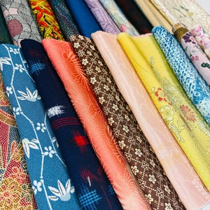 May include: A collection of folded fabric strips arranged in a row, featuring a variety of colors including blue, orange, yellow, brown, and pink, with diverse patterns such as floral, geometric, and abstract designs in various textures.