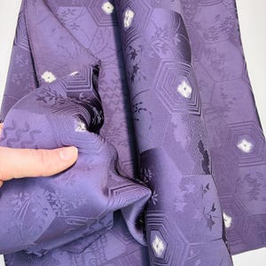 Op de afbeelding: Een gevouwen lengte paarse stof met een herhalend patroon van zeshoeken, bloemmotieven en geometrische lijnen. Het textiel heeft een gladde, satijnachtige afwerking met kleine, witte, stervormige accenten verspreid over het ontwerp.