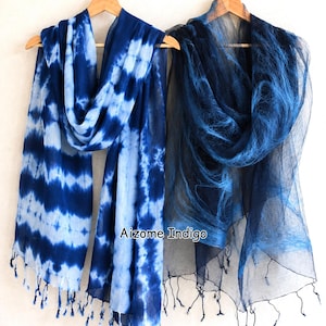 Può includere: Due sciarpe tinte con indaco appese a grucce di legno. La sciarpa a sinistra ha un motivo tie-dye nelle tonalità del blu e del bianco, con frange alle estremità. La sciarpa a destra è blu scuro tinta unita con una trama trasparente. Il testo "Aizome Indigo" è visibile.