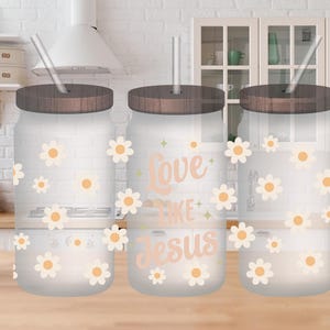 Puede incluir: Tres vasos de vidrio transparente con tapas marrones y pajitas. Los vasos tienen un patrón de margarita blanca y el vaso del medio tiene el texto "Love Like Jesus" en rosa.