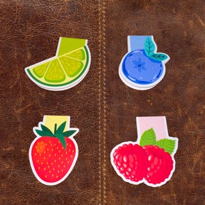 May include: Four colorful magnetic bookmarks featuring a lime wedge, a blueberry, a strawberry, and two raspberries. The bookmarks are on a brown leather background.