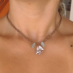 Puede incluir: Un collar de cordón marrón con un pequeño colgante de concha y dos cuentas de vidrio azul claro.