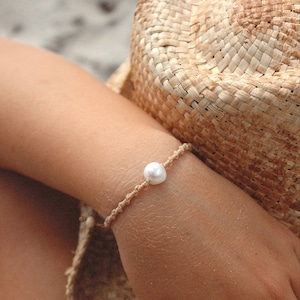 Può includere: Un semplice braccialetto con un'unica perla bianca su un cordoncino marrone. Il braccialetto è indossato al polso.