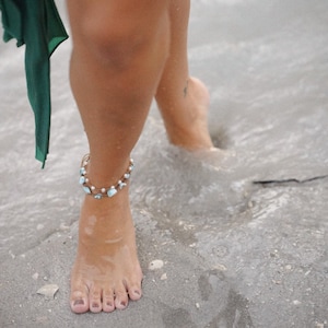 May include: A woman's bare foot with a delicate anklet made of small blue and white stones. The anklet is worn on the left ankle and the foot is resting on a sandy surface.
