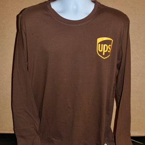 以下が含まれることがあります： 左胸に金のUPSロゴが付いた茶色の長袖Tシャツです。