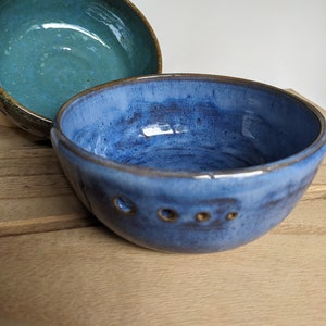 May include: Two handmade ceramic bowls. The front bowl is a deep blue with a glossy finish and three small holes near the rim. The second bowl is a teal color with a brown rim. Both bowls are on a wooden surface.