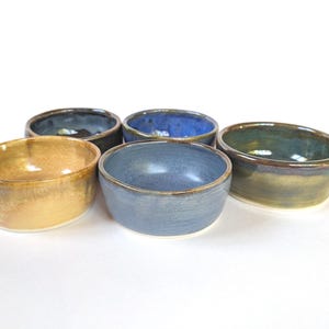 May include: Five handmade ceramic bowls in various colors: gold, blue, and green. Each bowl has a unique glaze and a rounded shape. The bowls are arranged on a white surface, showcasing their individual colors and textures.