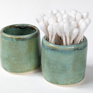May include: Two cylindrical ceramic containers with a glossy, mottled green glaze and dark blue-green rim accents. One container is empty, while the other is filled with a cluster of white cotton swabs with paper stems, standing upright.