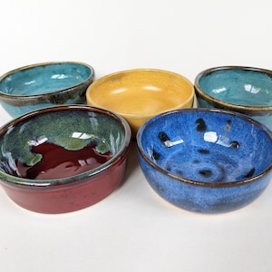 May include: A collection of five small, round ceramic bowls with varied glaze finishes. The set includes two light blue bowls, one mustard yellow bowl, one deep blue bowl, and one bowl featuring a dark red interior with a green rim.