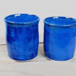 May include: Two cylindrical ceramic cups with a vibrant, deep blue glaze. Each cup features a smooth, glossy finish with subtle variations in color intensity across the surface, a slightly flared rim, and a clean, white base at the bottom.