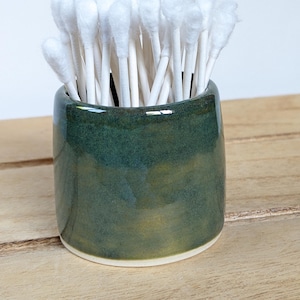 May include: A cylindrical ceramic holder with a glossy, mottled dark green and earthy yellow glaze. The container is filled with a cluster of white cotton swabs, each featuring a soft, rounded tip and a slender, straight white paper stem.