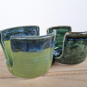 May include: Two handmade ceramic sponge holders in shades of green and blue. The holders have a curved design with a slot for a sponge. The glaze creates a textured, glossy finish. They are displayed on a wooden surface.