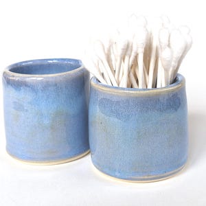 May include: Two cylindrical ceramic containers with a glossy blue glaze and a smooth finish. One container is empty, while the other is filled with a cluster of white cotton swabs. Both items feature a light-colored base rim and a simple design.