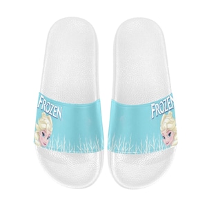 May include: A pair of white slip-on sandals with a blue and white Frozen design. The sandals feature a picture of Elsa from the movie Frozen and the word "Frozen" on each sandal.