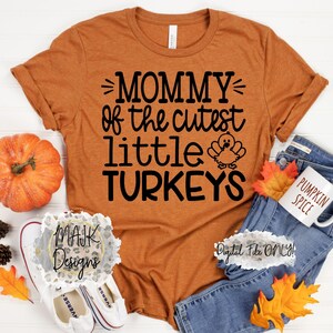 Może przedstawiać: Brązowa koszulka z napisem "Mommy of the cutest little turkeys" w kolorze czarnym. Obok tekstu znajduje się mały obrazek indyka. Koszulka leży na białym tle z dynią, dżinsami i kubkiem.