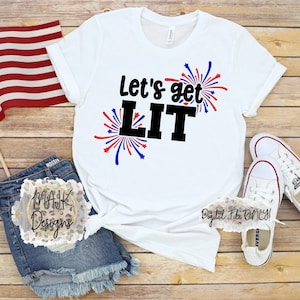 May include: White t-shirt with the text "Let's get LIT" in black with red and blue firework graphics.  The shirt is laying on a wood surface with a pair of white sneakers and a pair of denim shorts.