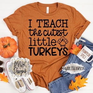 May include: An orange t-shirt with the text "I TEACH the cutest little TURKEYS" in black. A cartoon turkey is printed below the text. The shirt is displayed with a pumpkin, jeans, a mug that says "PUMPKIN SPICE", and fall leaves.