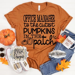 May include: A brown t-shirt with the text "Office Manager to the cutest Pumpkins in the patch" in black lettering. The shirt is lying on a white surface with a pumpkin, jeans, and a mug that says "Pumpkin Spice".