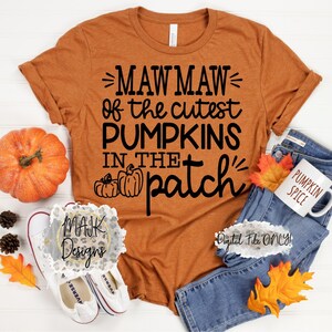 Könnte beinhalten: Ein braunes T-Shirt mit dem Text "Mawmaw of the cutest pumpkins in the patch" in schwarzer Schrift. Das Shirt wird auf einer weißen Oberfläche mit einem Kürbis, Herbstblättern und einem Paar weißen Turnschuhen gezeigt.