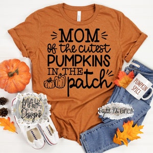 May include: A brown t-shirt with the text "MOM of the cutest PUMPKINS IN THE patch" in black. The shirt is laying on a white surface with a pumpkin, jeans, and a mug that says "PUMPKIN SPICE".