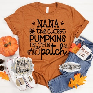 Könnte beinhalten: Orangefarbenes T-Shirt mit schwarzem Text, der "NANA of the cutest PUMPKINS in the patch" lautet, mit einer kleinen Grafik von drei Kürbissen. Das Shirt liegt auf einer weißen Oberfläche mit einem Paar weißen Turnschuhen, einem Kürbis und einer blauen Jeans mit einer weißen Tasse, auf der "PUMPKIN SPICE" steht.