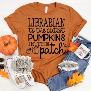May include: A brown t-shirt with the text "LIBRARIAN to the cutest PUMPKINS IN THE patch" in black lettering. The shirt is lying on a white surface with a pumpkin, pine cones, and autumn leaves.