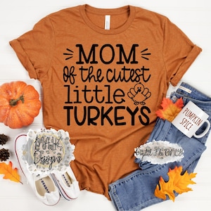 May include: An orange t-shirt with the text "MOM of the cutest little TURKEYS" in black. The image includes a pumpkin, a coffee mug that says "PUMPKIN SPICE", jeans, and fall leaves. The shirt is a digital file.