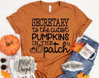 May include: A brown t-shirt with the text "SECRETARY to the cutest PUMPKINS IN THE patch" in black lettering. The shirt is laying on a white surface with a pumpkin, jeans, and a mug that says "PUMPKIN SPICE".