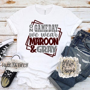 May include: White t-shirt with the text "On Gameday We Wear Maroon & Gray" in maroon and gray lettering inside a gray and maroon square frame. The shirt is laying on a wood surface with a pair of black converse shoes and a pair of blue jeans.