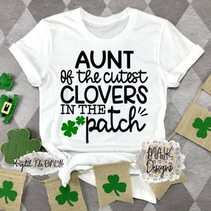 May include: White t-shirt with black text that reads "Aunt of the cutest clovers in the *patch*" with a four-leaf clover graphic.  The shirt is displayed on a gray and white checkered background with green shamrock decorations.