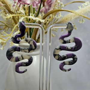 Op de afbeelding: Een paar paarse en witte slangen oorbellen. De oorbellen zijn gemaakt van polymer clay en hebben een textuur.