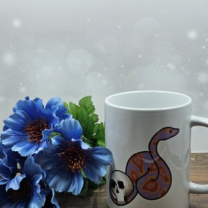 May include: White ceramic mug with a graphic of a purple and orange snake wrapped around a skull. The mug is sitting on a wooden surface with blue flowers in the foreground.