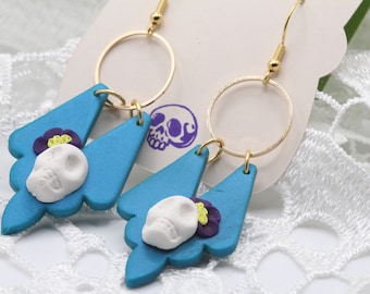 May include: A pair of blue and white dangle earrings with a skull and flower design. The earrings are made of polymer clay and have gold hooks.